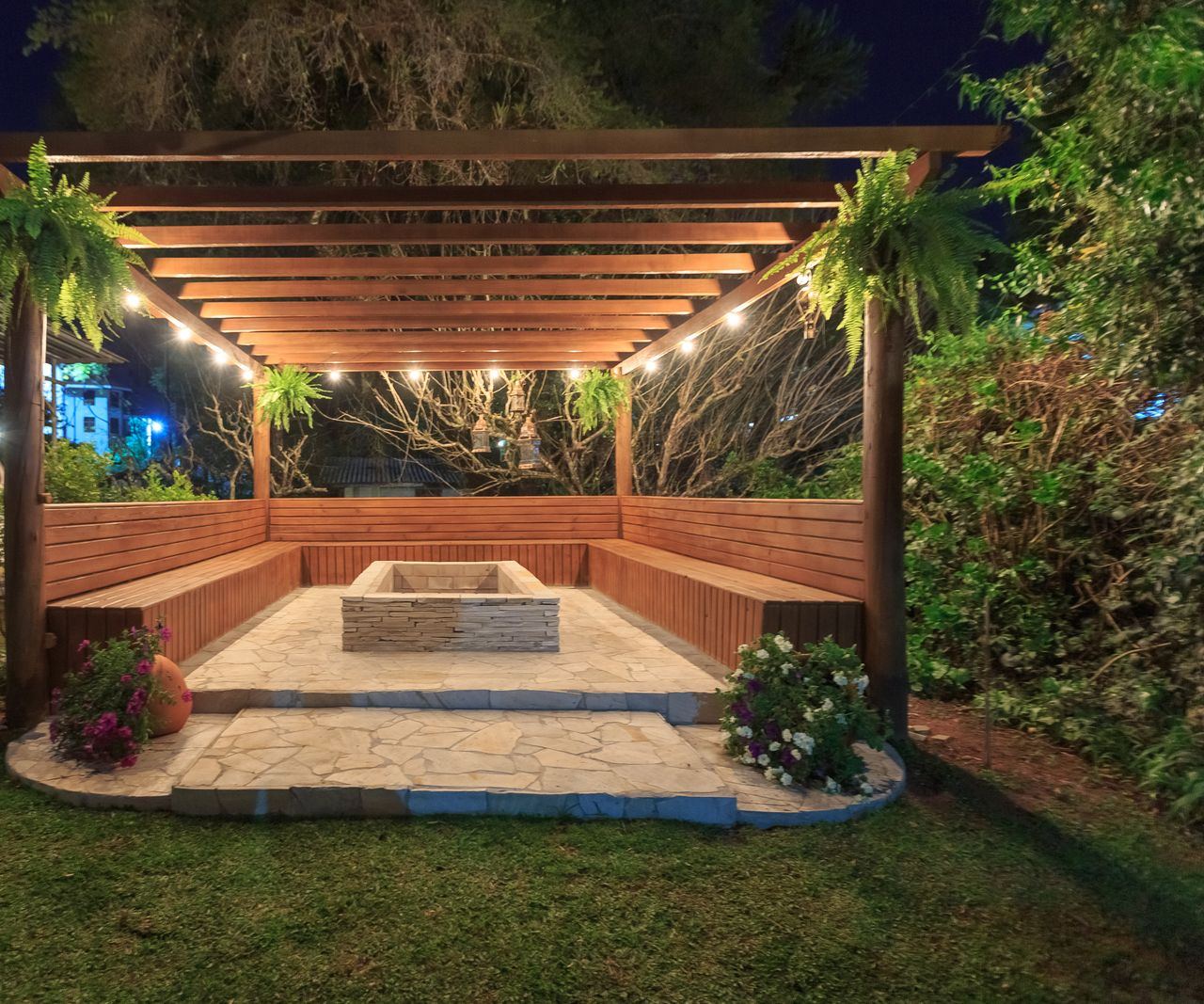 A wooden pergola with string lights over built-in benches and a stone fire pit at dusk